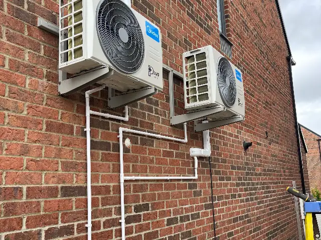 Two outdoor air conditioning units wall-mounted on brackets on the side of a brick house
